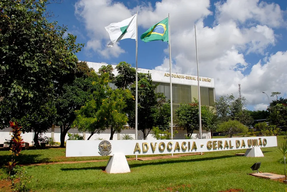 Prédio da Advocacia-Geral da União com bandeira do Brasil e paisagem verdejante ao redor.